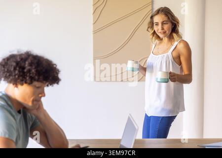 Couple at dining table, woman in sleeveless white top holding two ceramic mugs, man working laptop Stock Photo
