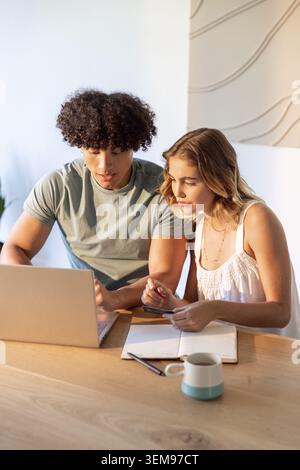 Couple wearing muted green tee and white top, sitting in home workspace using laptop and smartphone Stock Photo