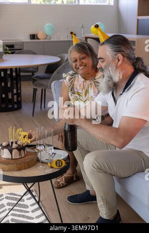 Couple with mature man and senior woman celebrating birthday on gray sofa, holding bottle near cake Stock Photo
