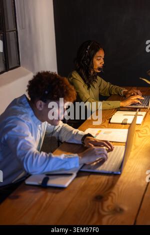 Diverse coworkers working at wooden desk in office wearing headsets and typing on laptops Stock Photo