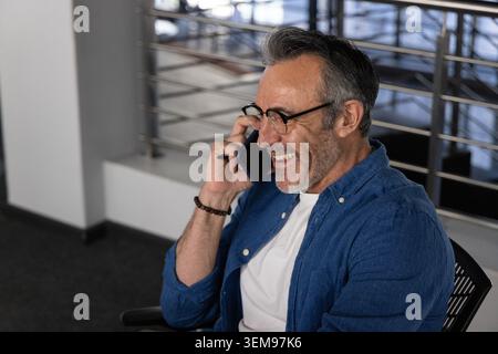 Middle-aged man sitting in office smiling while talking on smartphone, wearing glasses and bracelet Stock Photo