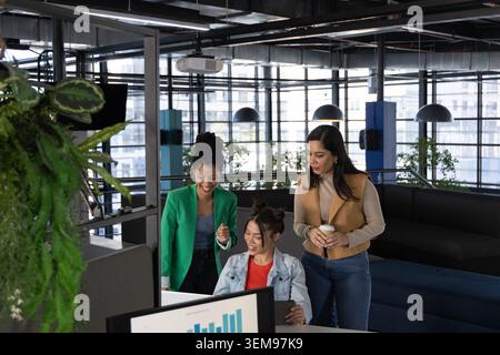 Diverse female colleagues analyzing charts on monitor at modern open-plan office with coffee cup Stock Photo