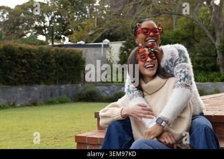 Diverse female friends sitting on brick steps in backyard, wearing wreath and reindeer glasses. Festive, eyewear, couple, outdoor, garden, playful, co Stock Photo