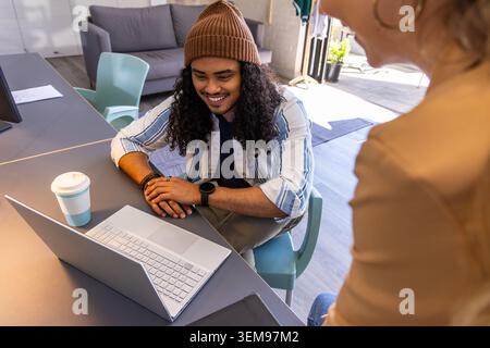 Diverse coworkers leaning and collaborating at coworking table, man wearing beanie, laptop, coffee. Collaboration, teamwork, sunlit, modern, office, s Stock Photo