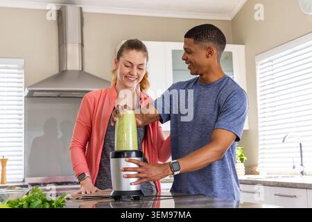Diverse couple in sportswear making green smoothie at kitchen island using blender and smartwatches. Duo, athleticwear, home, countertop, granite, her Stock Photo