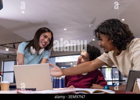 Diverse coworkers leaning in, collaborating at coworking table with open laptop and headphones Stock Photo
