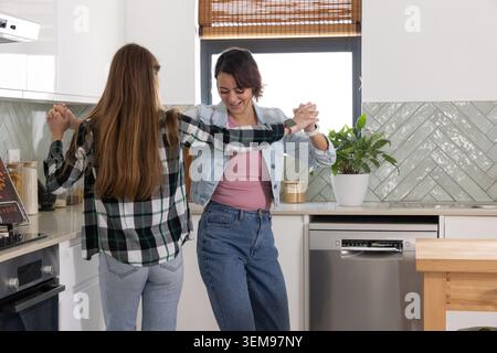 Two women holding hands and dancing in bright home kitchen by island with potted plant Stock Photo