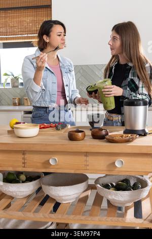 Female friends standing and preparing green smoothie in home kitchen with blender jar and avocados Stock Photo