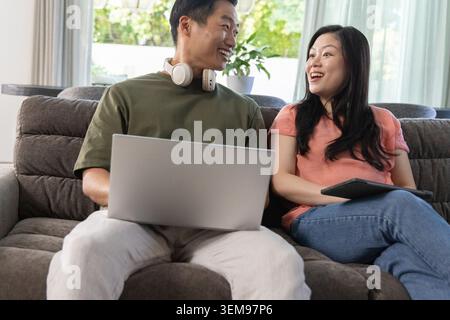Asian couple sitting on plush couch talking, smiling while using laptop and tablet with headphones Stock Photo