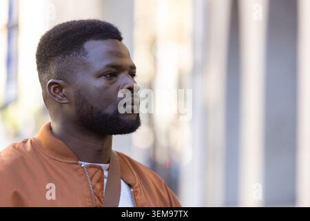 Man standing on city sidewalk wearing rust-orange zip-up jacket with brown crossbody strap Stock Photo