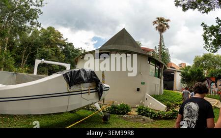 A round concrete structure shows partial collapse and debris around the curved facade. Nearby greenery, trees, and a pale sky frame the scene while a Stock Photo
