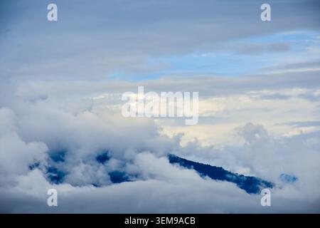 A dramatic ridge rises through thick clouds and mist, with patches of blue sky breaking the overcast, evoking serenity and grandeur in high-altitude n Stock Photo
