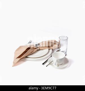 Sophisticated table place setting featuring stacked white ceramic plates, folded beige linen napkin with decorative ring, polished silverware, coffee Stock Photo