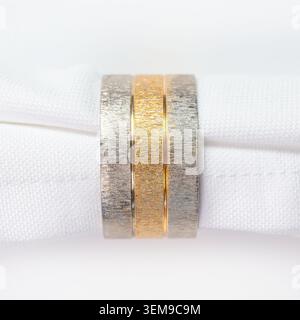 Set of three elegant brushed metal napkin rings featuring two silver and one gold tone displayed on crisp white linen. Stock Photo