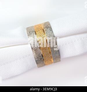 Close-up of three elegant textured wedding bands resting on a soft white fabric napkin. Two matte silver rings with brushed finish beautifully frame a Stock Photo