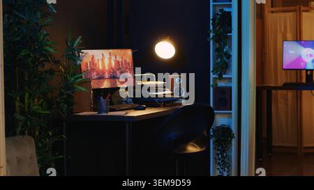 Work desk with computer and house plants in empty house interior with warm lighting. Living room illuminated by neon lights with 3D rendered animations running on powerful PC monitors Stock Photo