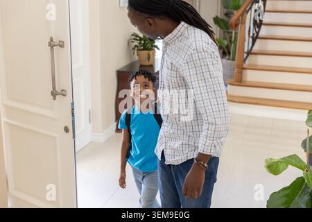 Male guardian and school-aged boy standing in home foyer holding backpack beside open front door Stock Photo