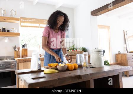 African American woman slicing fresh fruit with chef's knife on wooden island in farmhouse kitchen Stock Photo