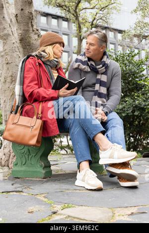 Senior couple sitting on bench in urban courtyard, woman wearing red coat reading black notebook Stock Photo