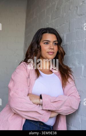 Female leaning with arms crossed by brick wall in corner, wearing pink top and smartwatch Stock Photo