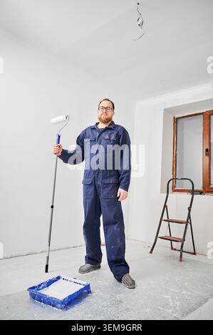 Worker holding paint roller on extension pole standing in bright room, looking at camera. Interior renovation stage with white walls, finishing work and home improvement concept Stock Photo