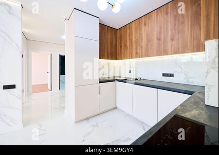 Modern corner kitchen with white cabinets and wooden upper units in bright apartment interior. Minimal design with marble walls, dark countertop and sleek contemporary layout concept Stock Photo