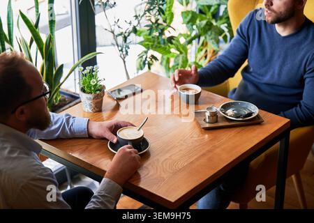 Two men having coffee conversation at cafe table, informal meeting or friendly chat, relaxed atmosphere with drinks, smartphone on table and green plants in bright interior Stock Photo