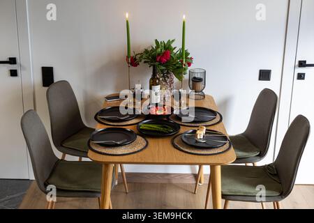 Candlelit dining table set for four. Modern home dining table with candles, wine, flowers, black plates, and vegetables prepared for an intimate Stock Photo