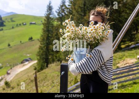Woman carrying daisies in mountain countryside. Woman in sunglasses and striped sweater holds a metal bucket of fresh daisies on a sunny green Stock Photo