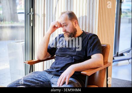 Concerned white bearded man looking down in thoughtful pose, contemplating decisions and next steps while seated in lounge, subdued expression and casual surroundings suggest planning, uncertainty, Stock Photo
