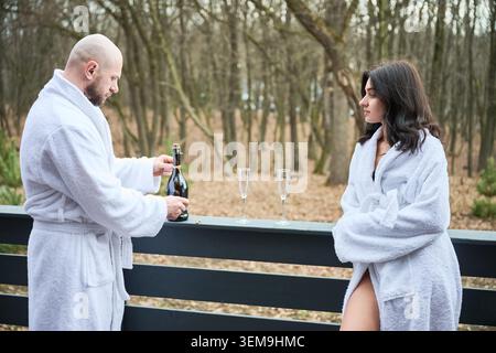 Man opening champagne bottle on terrace while woman watches in bathrobe. Outdoor spa retreat, forest background, romantic moment, relaxation, luxury vacation and wellness concept Stock Photo
