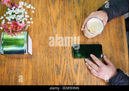 Woman using smartphone and holding latte at outdoor cafe, top view of hands on wooden table with flowers and napkin holder, relaxed leisure moment in daylight Stock Photo