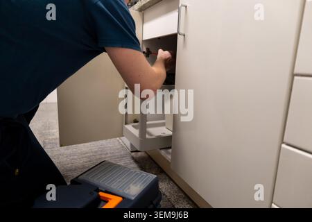 Person crouching, repairing a kitchen cabinet door using a screwdriver Stock Photo