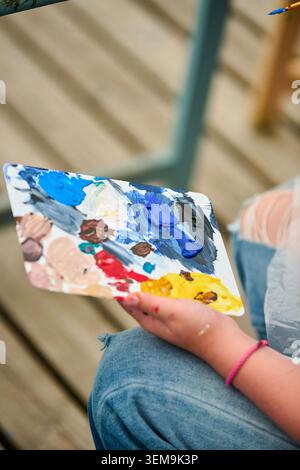 Close up of artist hand holding palette filled with vibrant oil paints including blue, yellow, red, and white shades. Wooden deck background and casua Stock Photo