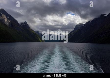 View from the stern of a cruise ship leaving a foaming wake on the dark waters of Geirangerfjord, flanked by towering green mountains with snow-capped Stock Photo
