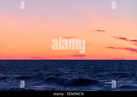 Calm deep blue ocean waves gently roll beneath gradient sunset sky of soft peach and pink hues, creating peaceful and serene coastal atmosphere Stock Photo