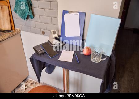 Compact kitchen table setup displaying smartphone, clipboard with blank paper, blue folder, notepad, pen, red apple, and empty glass cup against white Stock Photo
