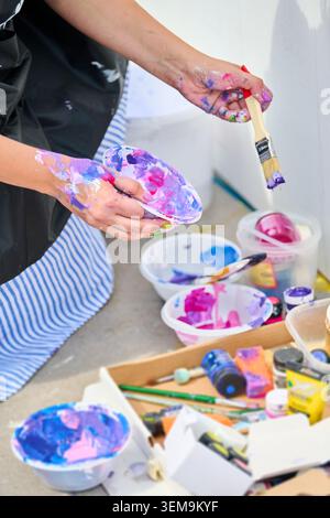 Artists hands covered in mixed paint holding brush and palette bowl with vibrant pink and purple hues, surrounded by various mixing bowls filled with Stock Photo
