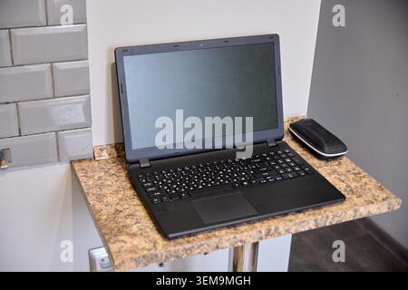 Open black laptop with dark screen positioned on small granite surface against plain white wall with gray subway tiles on side. Wireless mouse placed Stock Photo