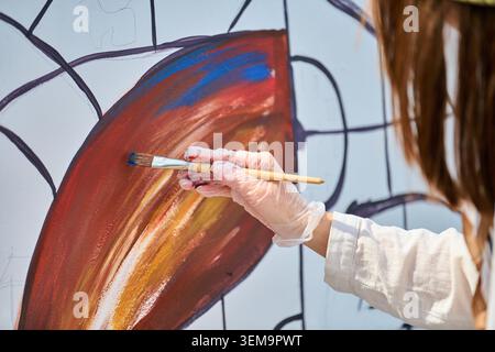 Artist wearing white glove applies brown and blue paint to abstract mural design on white canvas using paintbrush in bright environment. Focus on hand Stock Photo