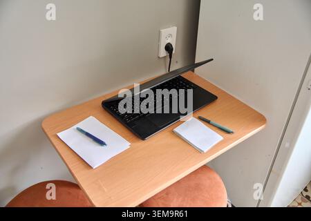 Compact wooden desk holding partially closed black laptop with two notebooks and pens arranged on either side, set against light wall with power outle Stock Photo