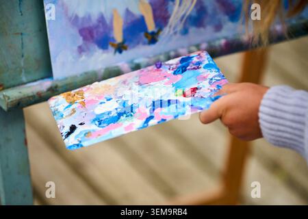 Close up view of hand holding painters palette with mixed bright colors near canvas featuring purple and blue tones. Soft daylight illuminates creativ Stock Photo