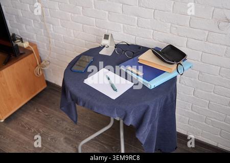 Compact table draped with blue fabric displays pen on blank paper, eyeglasses, digital clock, and stacked books under soft indoor lighting near white Stock Photo
