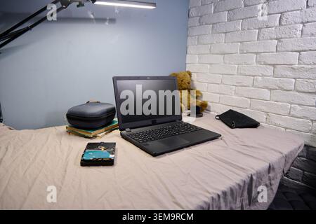 Black laptop positioned on beige bed near white brick wall with stacked books, teddy bear, wallet, and desk lamp creating organized and comfortable wo Stock Photo