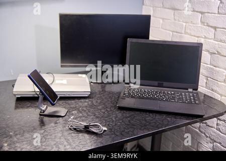Workspace featuring closed laptop under widescreen monitor, open laptop with black keyboard, smartphone on stand, white brick background, soft natural Stock Photo