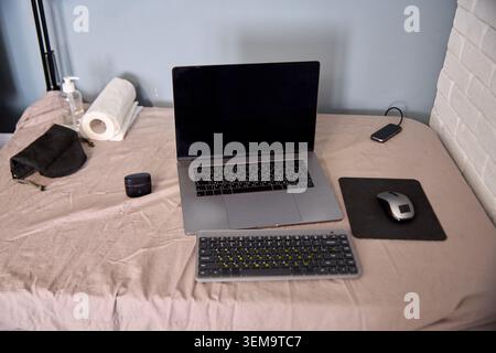 Modern workspace featuring closed laptop with external keyboard and wireless mouse on mousepad beside roll of paper towels hand sanitizer and black fa Stock Photo