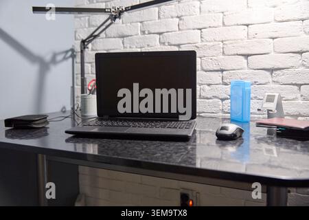 Black laptop computer with closed screen sits on glossy black marble desk alongside mouse, blue water bottle, and small clock, illuminated by directio Stock Photo
