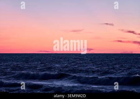 Deep blue ocean waves gently roll under colorful gradient sky transitioning from pink to purple during sunset with soft scattered clouds on horizon Stock Photo