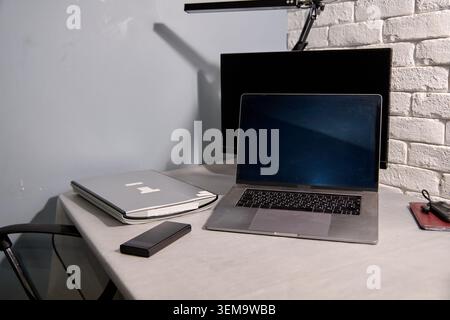 Minimalist office desk setup featuring open laptop, closed silver notebook, external black monitor, and adjustable lamp against textured white brick w Stock Photo