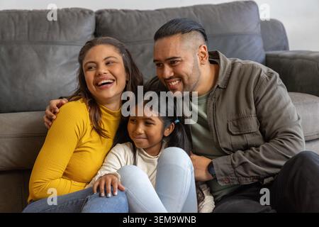 Asian family sitting close on gray sofa in living room, parents embracing daughter in white sweater Stock Photo